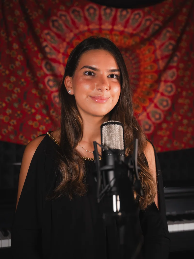 Portrait of Sirine in her studio beside a microphone, with a rich red tapestry behind her.